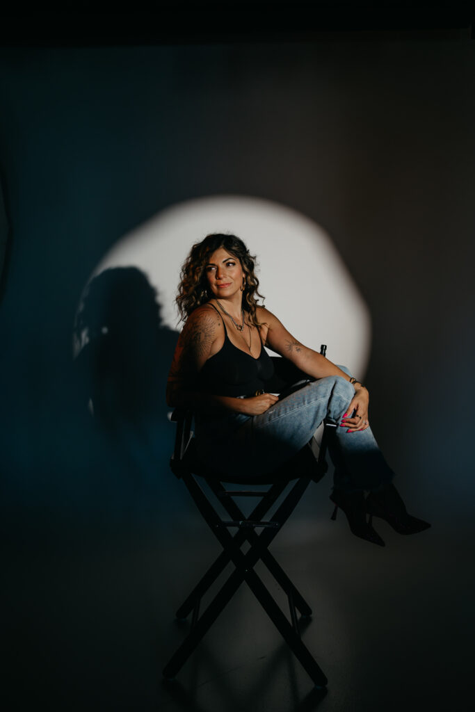 Dramatic studio portrait of Dr. Lauryn in black attire sitting in a director’s chair under a spotlight for her editorial brand session.