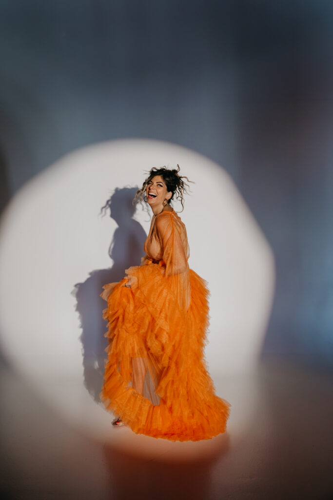 Dr. Lauryn in orange tulle gown sitting on the studio floor, smiling under a spotlight during her luxury brand photoshoot with Minneapolis photographer Mycah Bain.