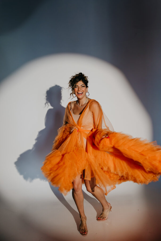 Dr. Lauryn in orange tulle gown sitting on the studio floor, smiling under a spotlight during her luxury brand photoshoot with Minneapolis photographer Mycah Bain.