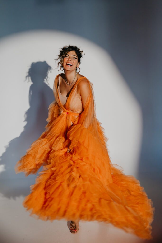 Dr. Lauryn in orange tulle gown sitting on the studio floor, smiling under a spotlight during her luxury brand photoshoot with Minneapolis photographer Mycah Bain.