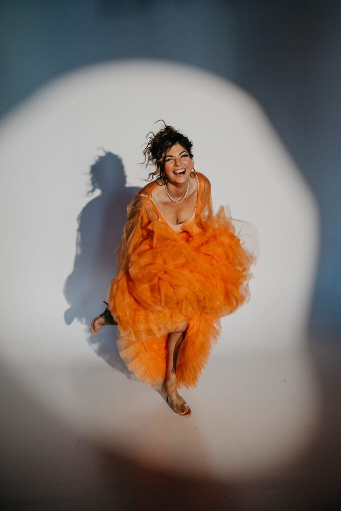 Dr. Lauryn in orange tulle gown sitting on the studio floor, smiling under a spotlight during her luxury brand photoshoot with Minneapolis photographer Mycah Bain.