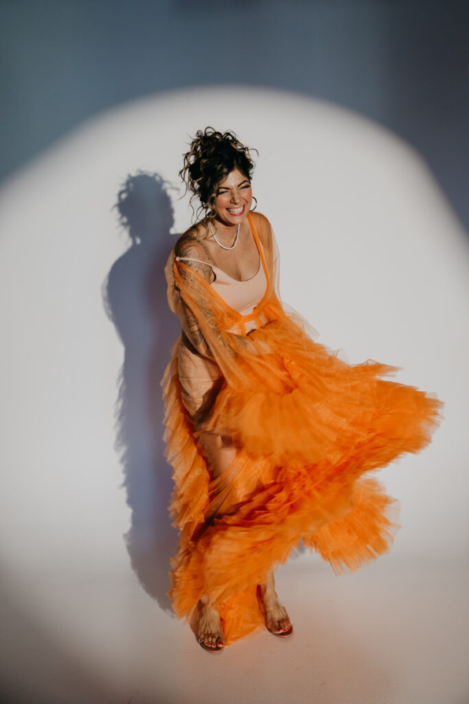 Dr. Lauryn in orange tulle gown sitting on the studio floor, smiling under a spotlight during her luxury brand photoshoot with Minneapolis photographer Mycah Bain.
