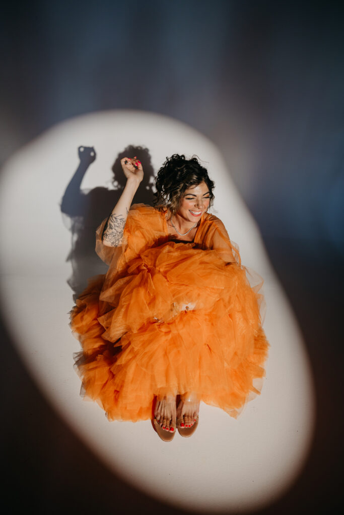 Dr. Lauryn in orange tulle gown sitting on the studio floor, smiling under a spotlight during her luxury brand photoshoot with Minneapolis photographer Mycah Bain.