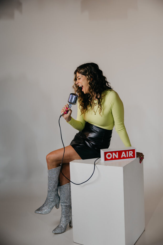 Playful image of Dr. Lauryn lying on the floor with glitter boots and microphone under an “On Air” sign during her brand photoshoot.