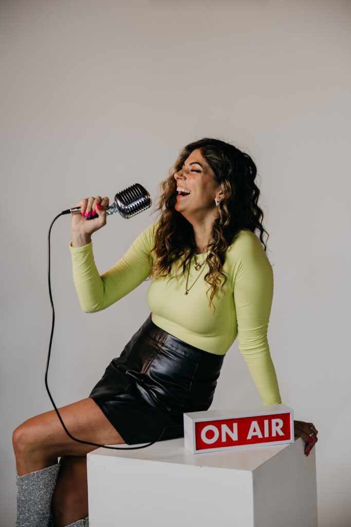 Playful image of Dr. Lauryn lying on the floor with glitter boots and microphone under an “On Air” sign during her brand photoshoot.