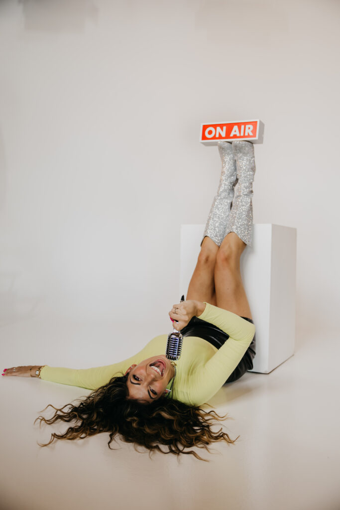 Playful image of Dr. Lauryn lying on the floor with glitter boots and microphone under an “On Air” sign during her brand photoshoot.