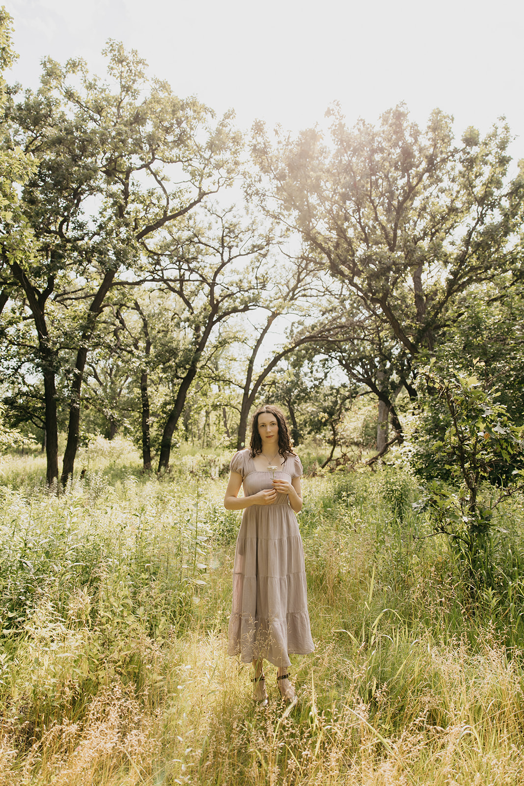 Senior girl standing in a sunlit meadow holding wildflowers, wearing a flowy neutral dress surrounded by tall grass and trees in Minneapolis.