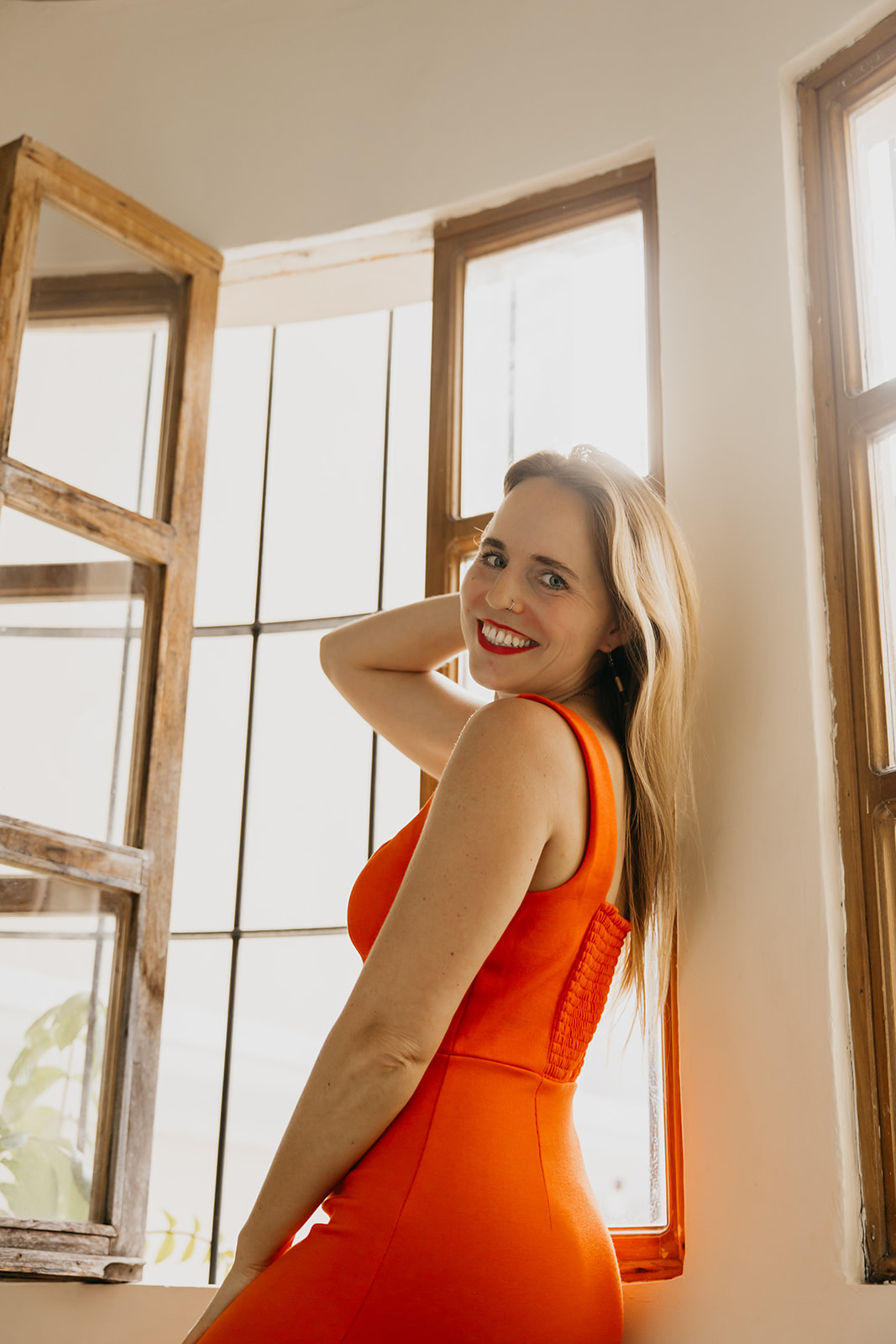 Creative entreprenuer standing by an open window wearing a vibrant orange dress, smiling confidently in natural light.