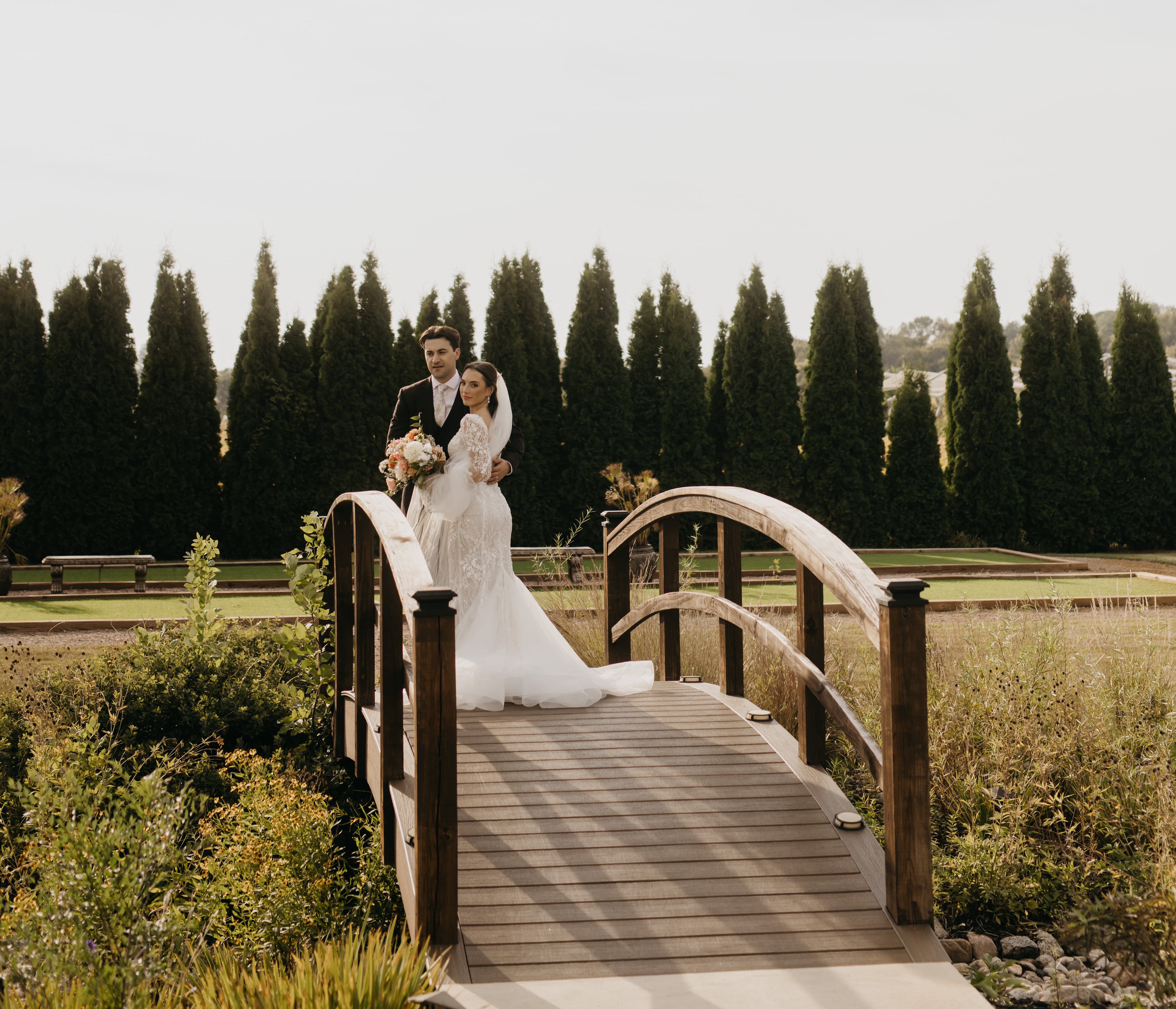 Bride and groom standing together on the wooden bridge at Ashery Lane Farm surrounded by greenery and tall pines.