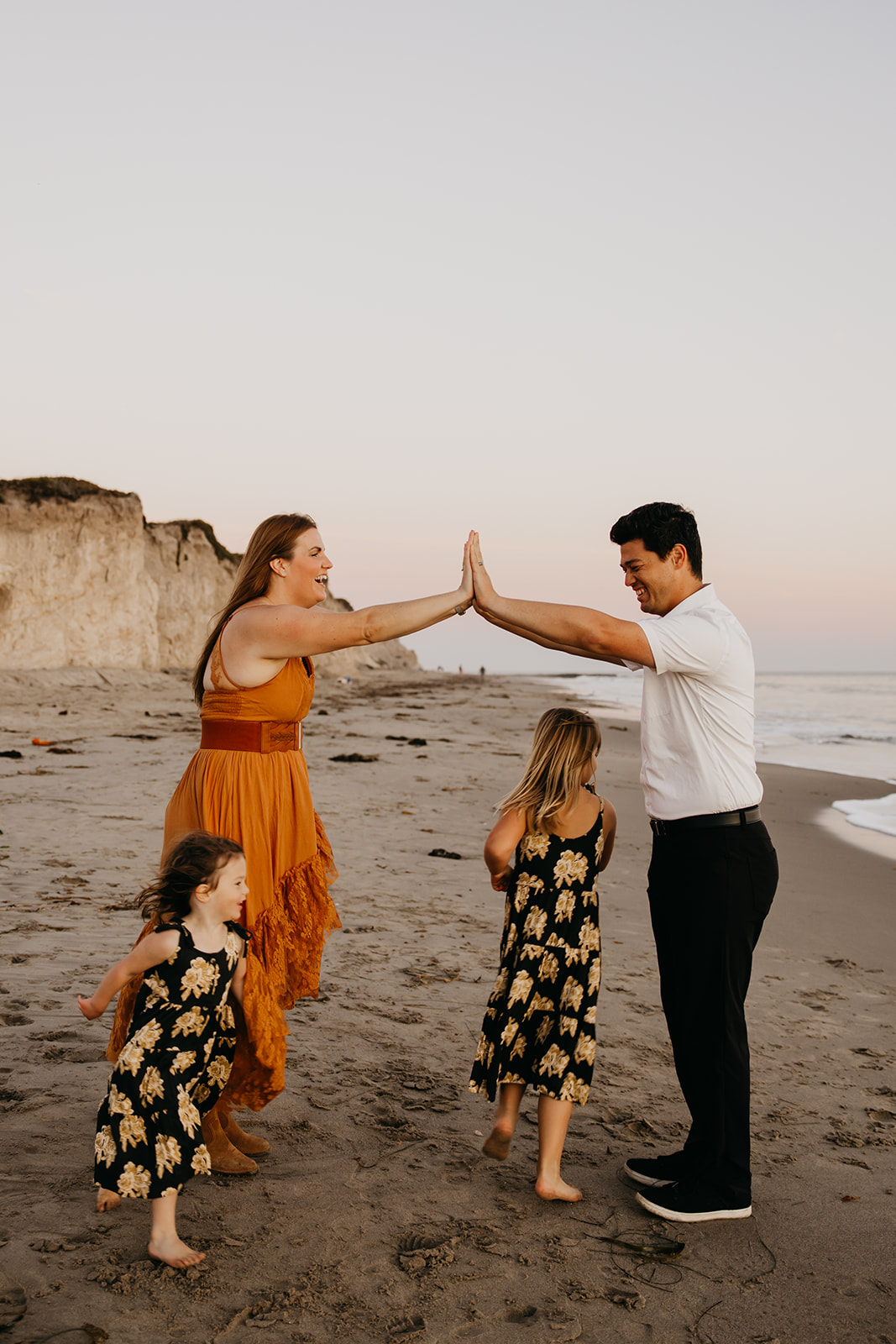 Family playing together on the beach during sunset, parents high-fiving as their daughters laugh and run around them.