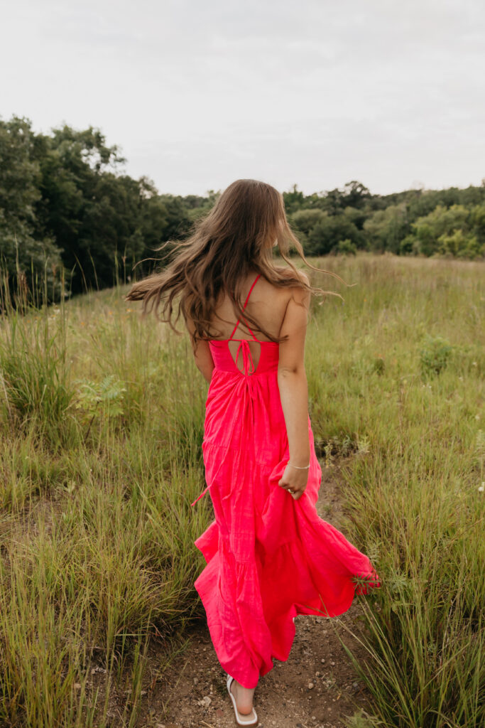 Senior girl walking through a field in a flowing bright pink dress, her hair blowing softly in the breeze with trees in the background.