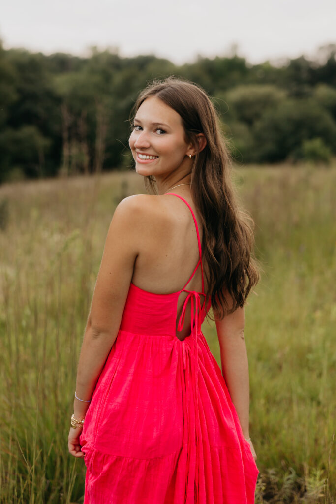 Senior girl smiling over her shoulder in a bright pink dress with open-back details, standing in a grassy field during golden hour.