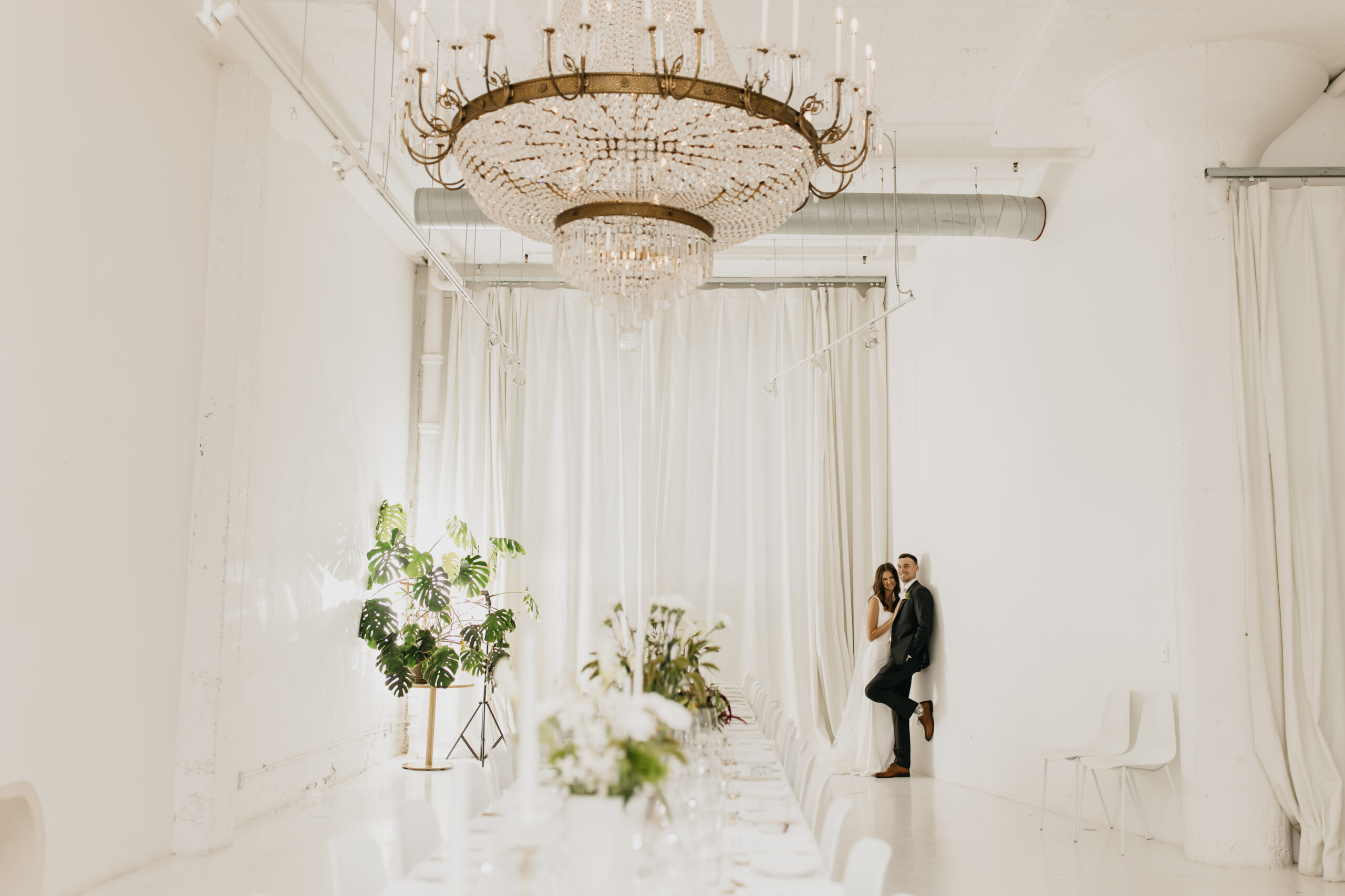 Bride and groom stand together at the end of a long, elegant dining table beneath a grand chandelier at their Paris Dining Club wedding in Minneapolis.