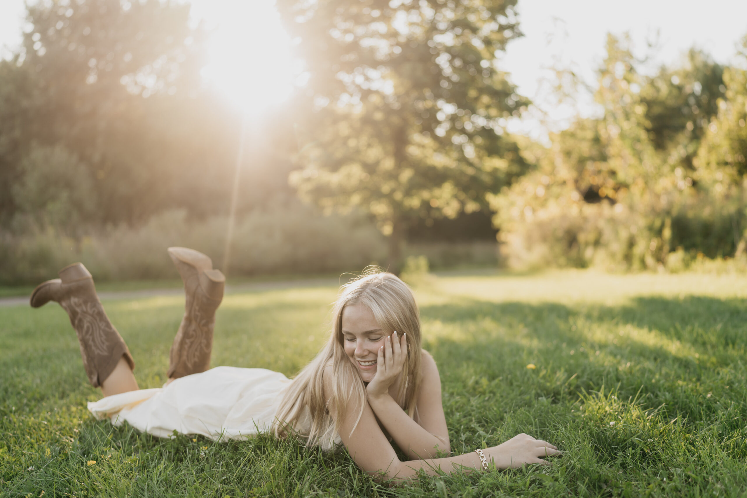 Senior girl lying in the grass at golden hour wearing a white dress and cowboy boots during a country-inspired senior photo session in the Twin Cities.