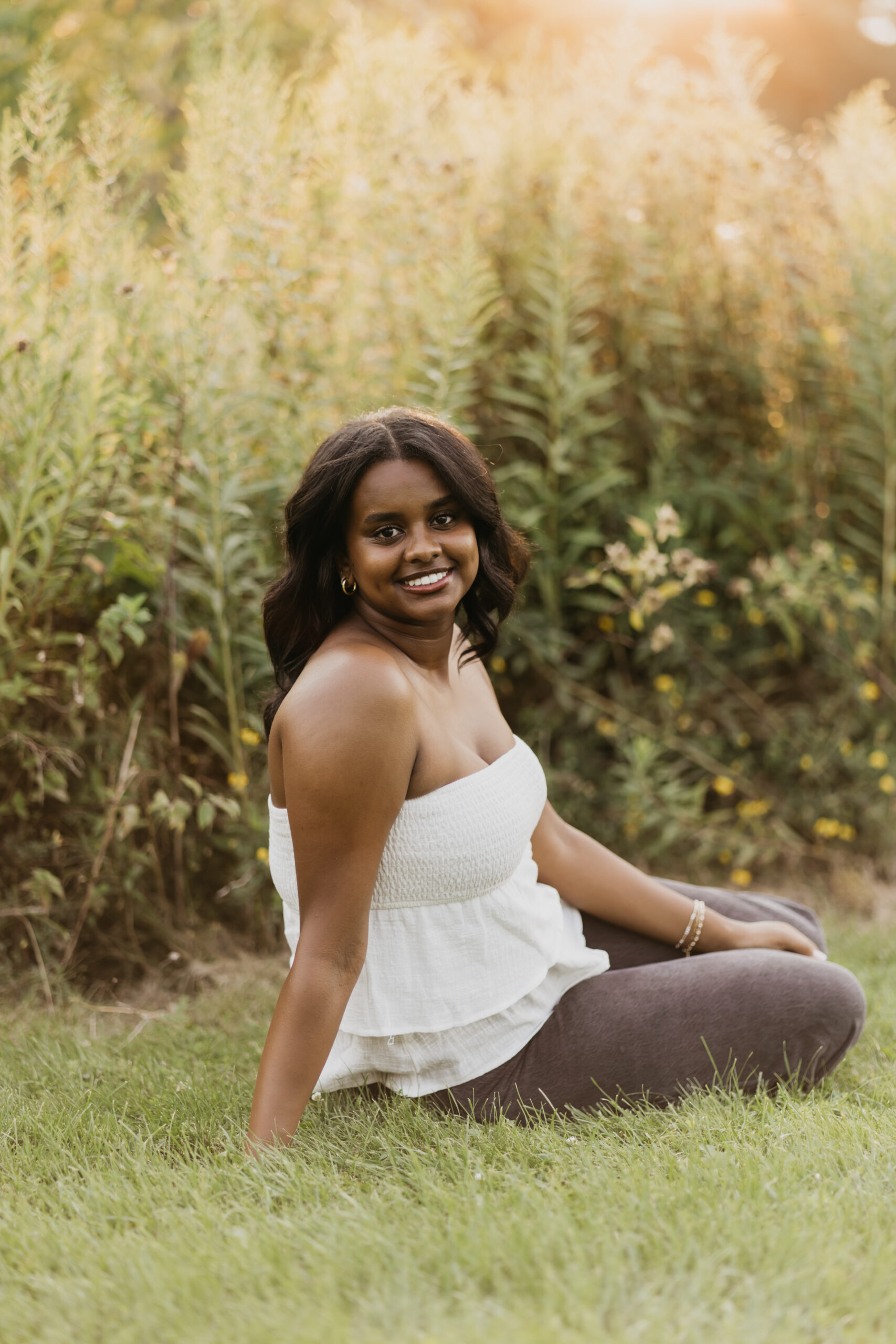 St Paul senior photo of a young woman sitting in the grass at golden hour, smiling confidently in a white strapless top and dark jeans surrounded by tall wildflowers.