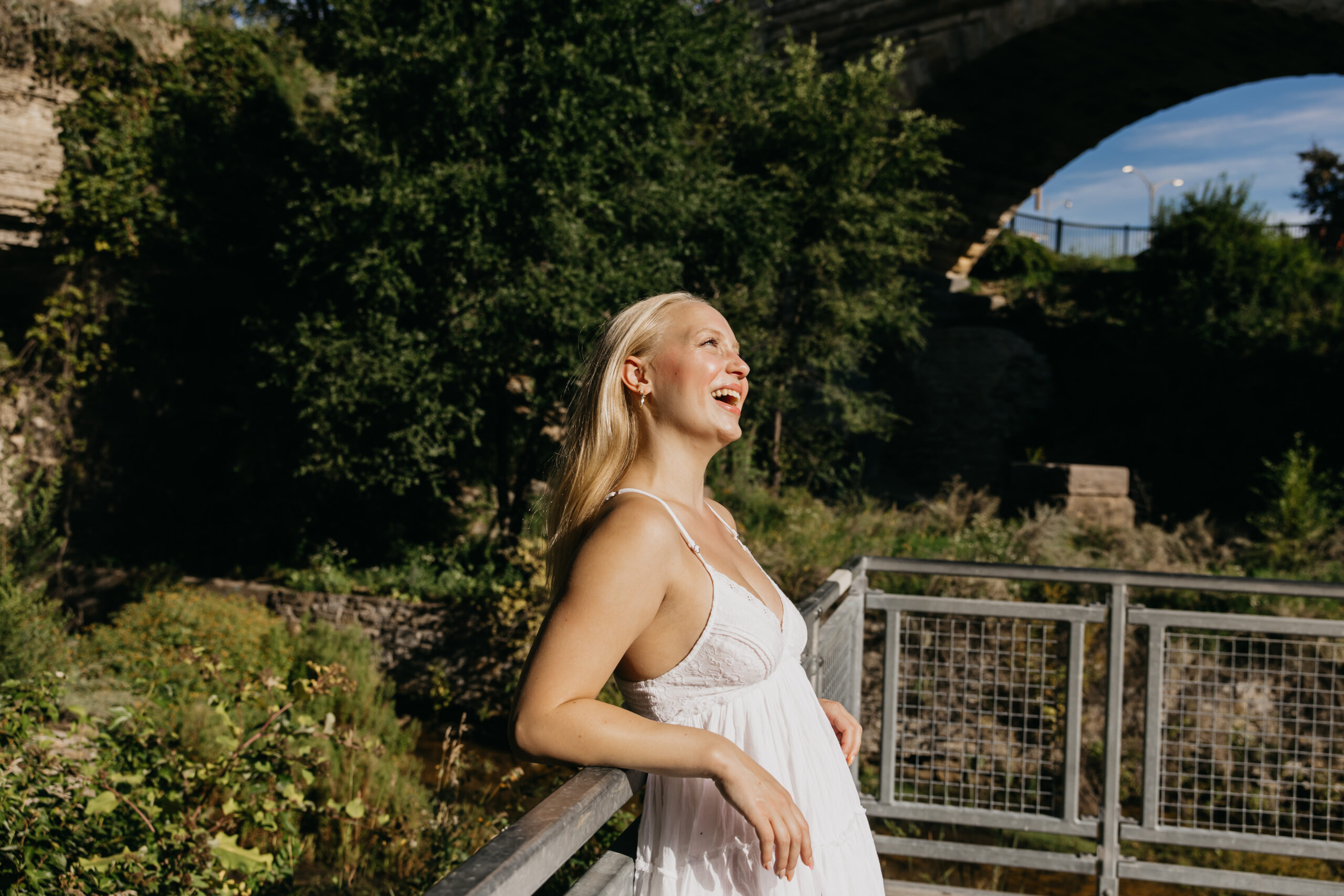 High school senior in a white dress laughing in the sunshine while leaning on a metal railing near a stone bridge during a Twin Cities senior photo session.