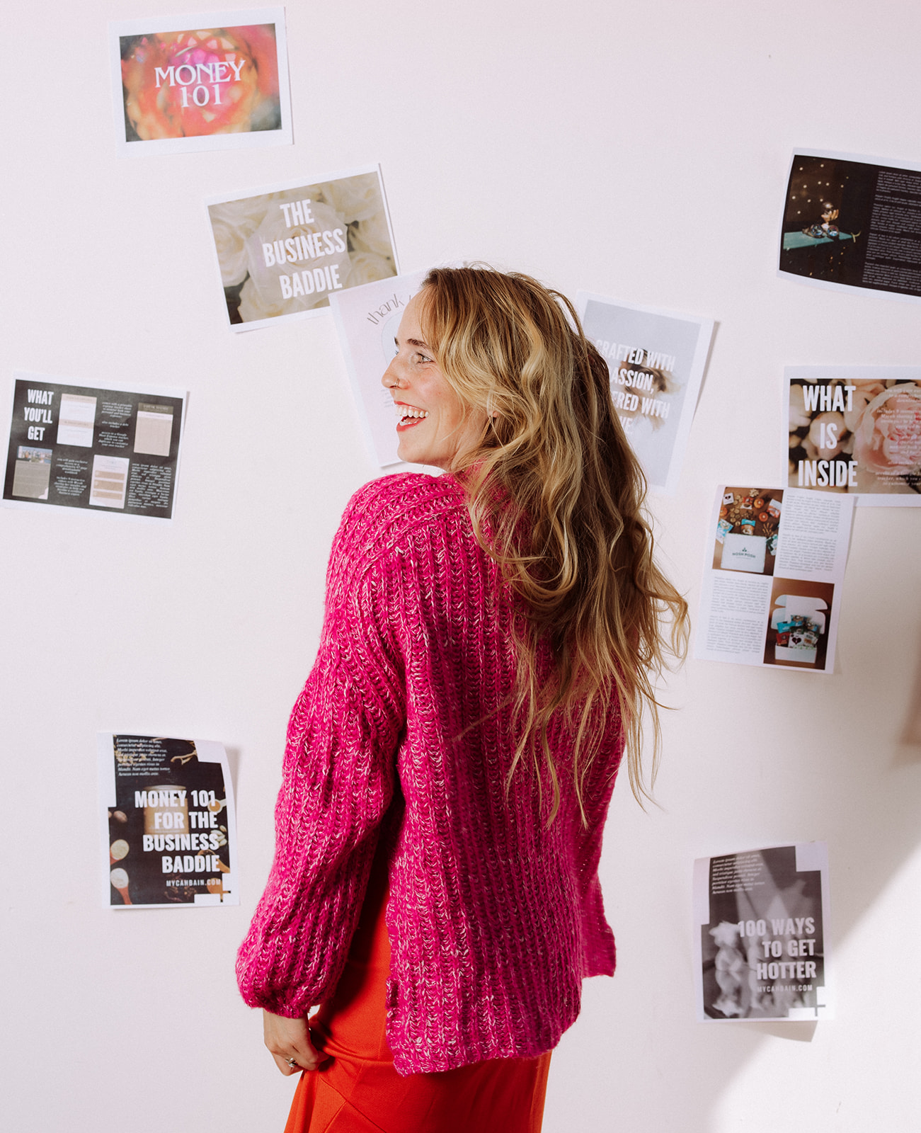 Woman in a bright pink sweater smiling while standing in front of a wall covered in business and money mindset inspiration boards, representing a money mindset for creative entrepreneurs.