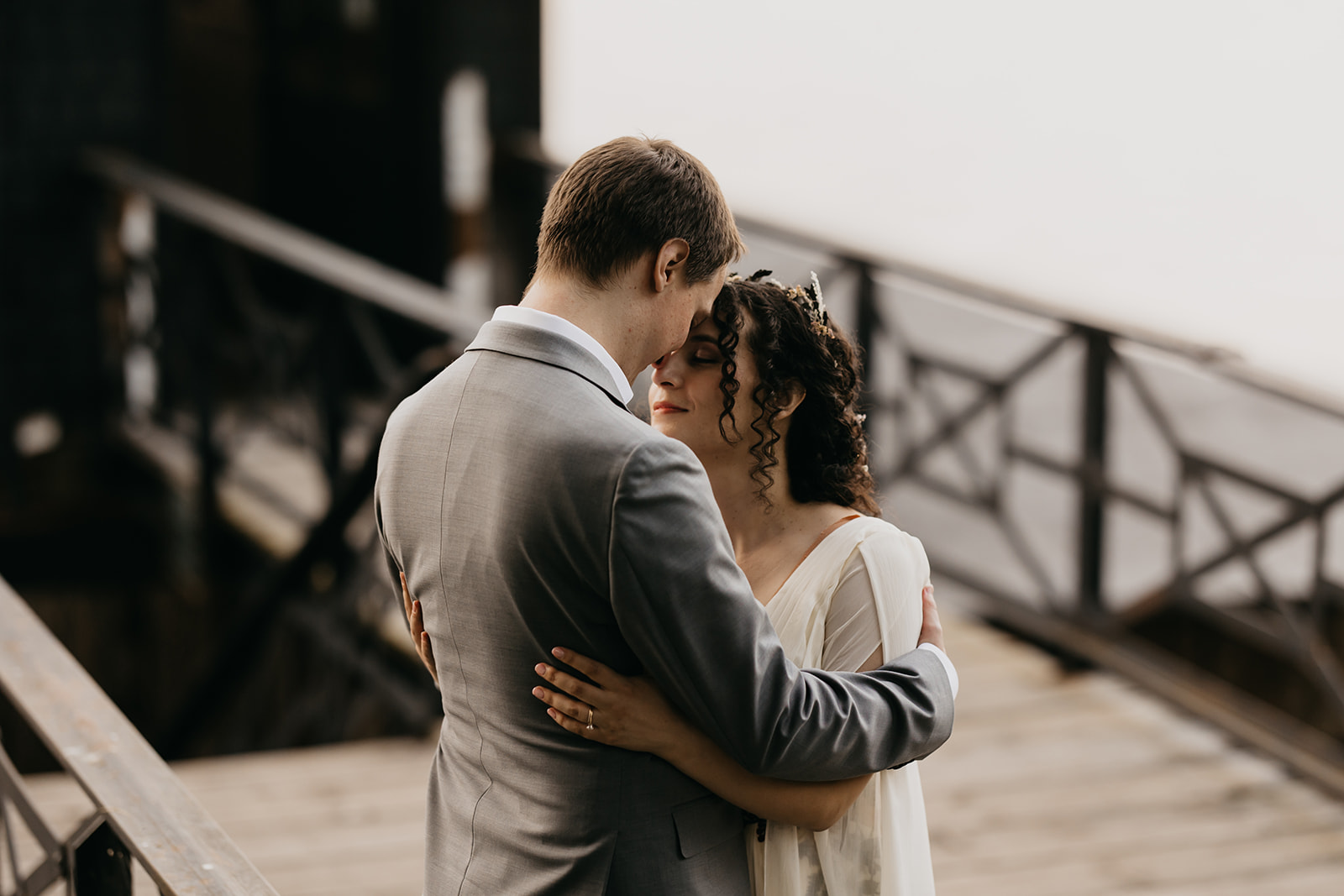 Bride and groom share an intimate moment on an industrial bridge, foreheads together, groom in gray suit holding bride in flowing white gown with floral crown, documentary wedding photography by Mycah Bain Photography Minneapolis