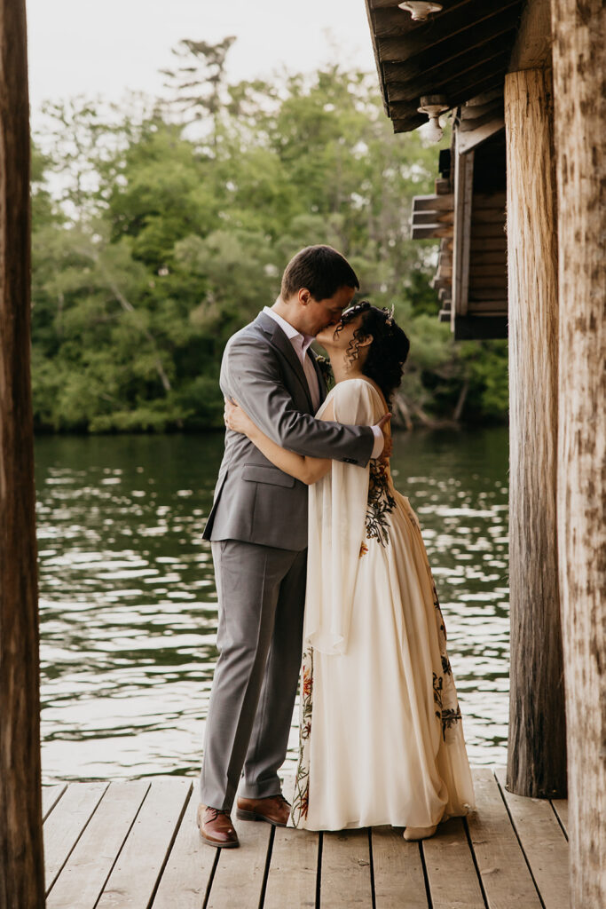 Artistic blurred foreground shot of bride and groom kissing on a lakeside dock surrounded by pine trees, intentional soft focus documentary wedding photography by Mycah Bain Photography Minnesota