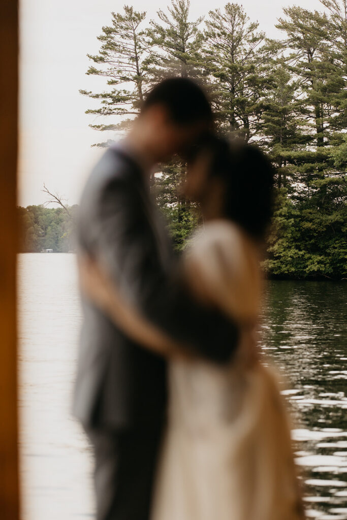 Bride in floral embroidered gown and groom in gray suit share first kiss on a rustic wooden boathouse dock by a Minnesota lake, framed by wooden posts and lush green trees, documentary wedding photography by Mycah Bain Photography