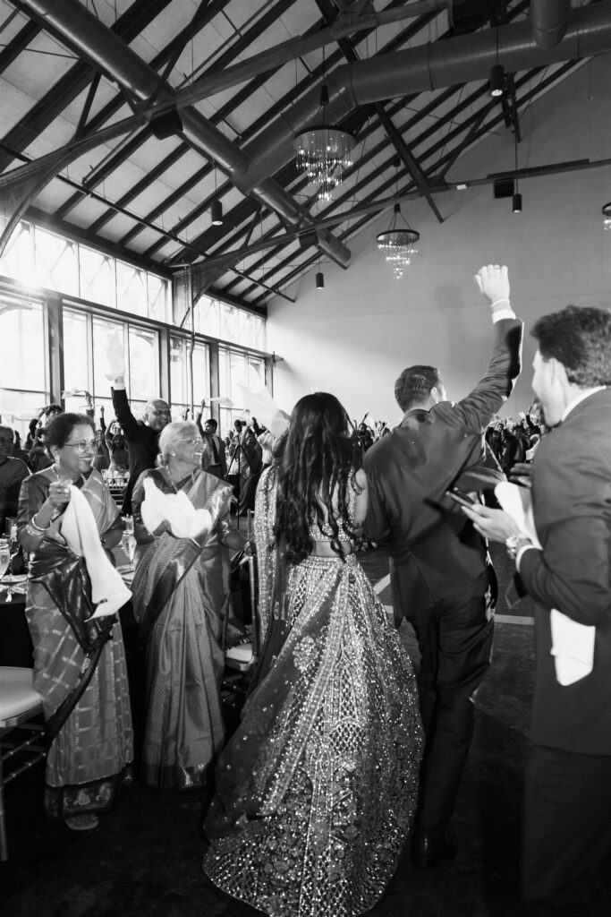 Black and white candid photo of Indian wedding reception in Minneapolis, bride in sequined lehenga facing cheering crowd with hands raised, grandmothers in saris celebrating in foreground, documentary wedding photography by Mycah Bain Photography