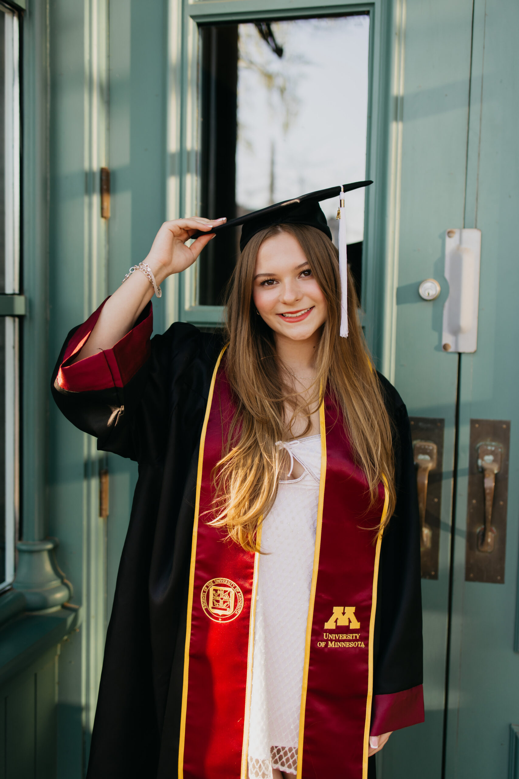College graduation photographer Minneapolis, University of Minnesota grad smiling in cap and gown at campus door, Mycah Bain Photography