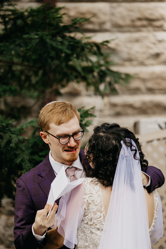 Bride embracing groom from behind during outdoor wedding first look, groom in purple shirt, bride in lace gown with veil, guests blurred in background, documentary wedding photography by Mycah Bain Photography Minneapolis
