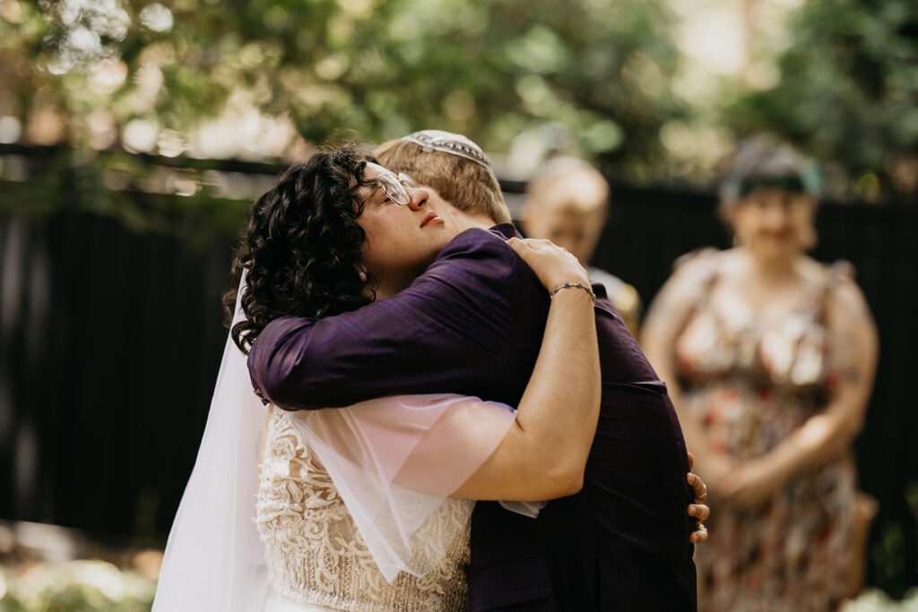 Groom in purple plaid suit with kippah holds bride in lace gown tightly against a stone wall backdrop during emotional first look at Jewish wedding in Minneapolis, documentary photography by Mycah Bain Photography
