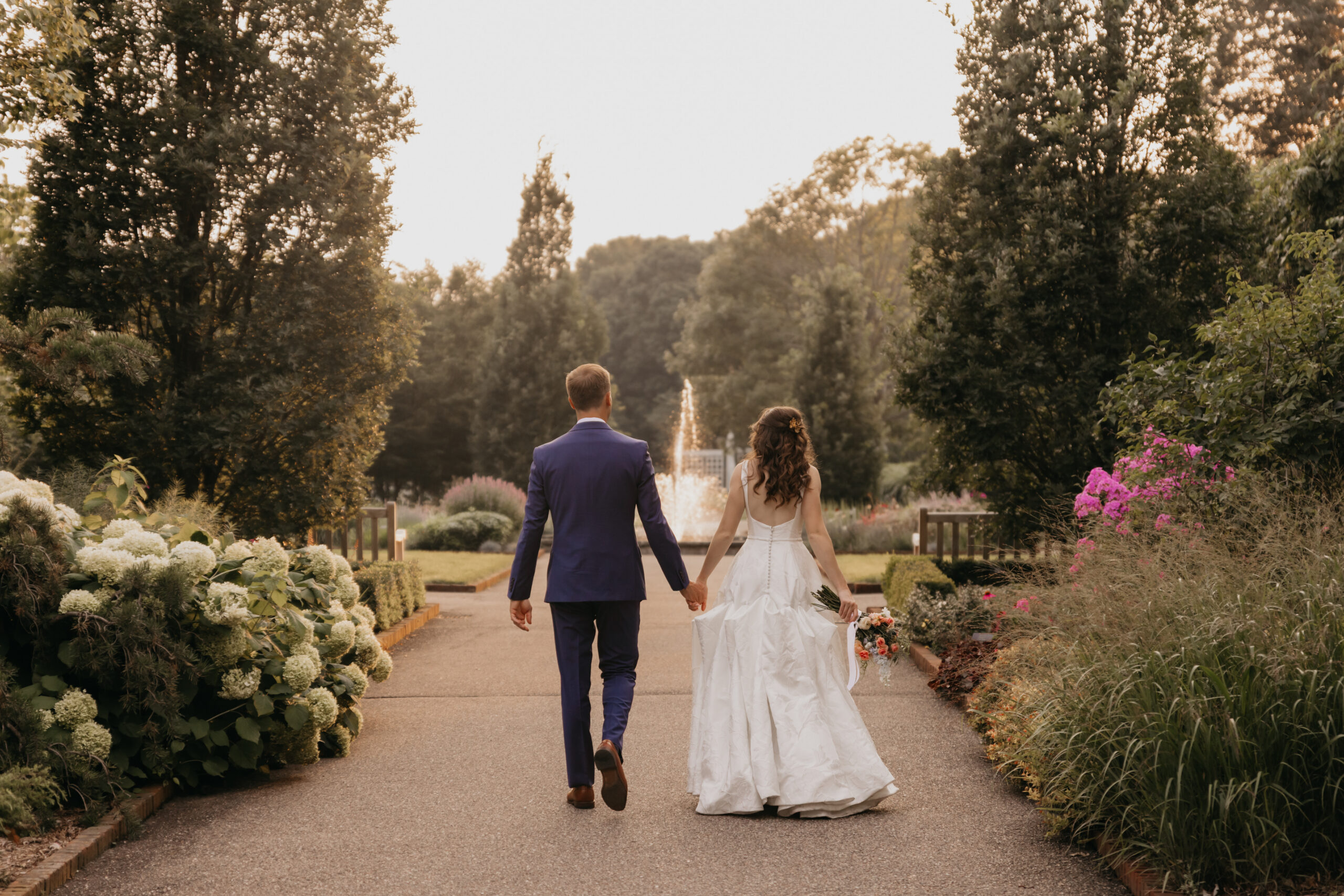 Bride and groom walking hand in hand through a lush garden path toward a glowing fountain at golden hour, captured by Mycah Bain Photography, Minneapolis wedding photographer