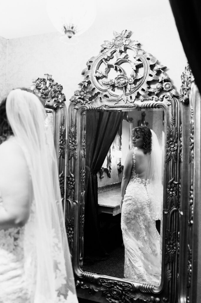 Black and white photo of bride in backless lace wedding gown and veil reflected in an ornate carved gold mirror while getting ready at Como Conservatory wedding in Saint Paul, documentary wedding photography by Mycah Bain Photography