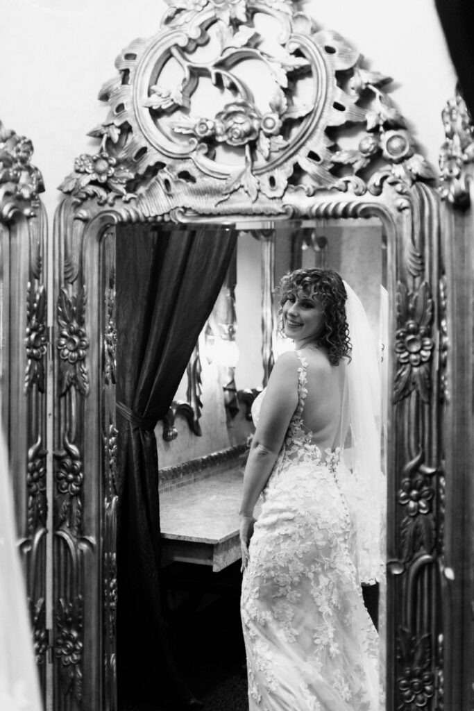  Black and white portrait of curly-haired bride in backless lace gown and veil smiling over her shoulder while reflected in an ornate carved mirror, Como Conservatory wedding Saint Paul, documentary photography by Mycah Bain Photography