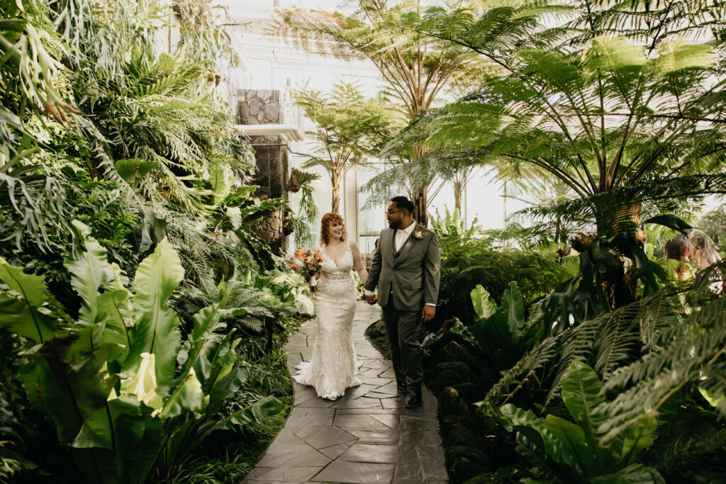 Bride in lace wedding gown holding colorful bouquet and groom in gray suit walk hand in hand through lush tropical greenhouse at Como Conservatory in Saint Paul, documentary wedding photography by Mycah Bain Photography
