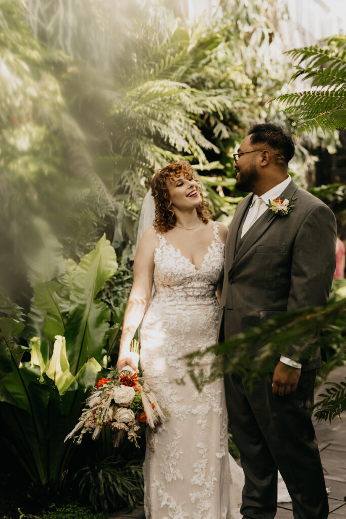 Bride with red curly hair in lace gown laughing with eyes closed holding wildflower bouquet as groom in gray suit smiles at her inside the tropical greenhouse at Como Conservatory Saint Paul, wedding photography by Mycah Bain Photography
