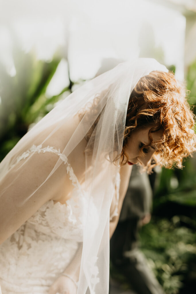 Close-up artistic portrait of red-haired bride with veil draped over her head bowing forward in soft backlit greenhouse light at Como Conservatory Saint Paul, documentary wedding photography by Mycah Bain Photography
