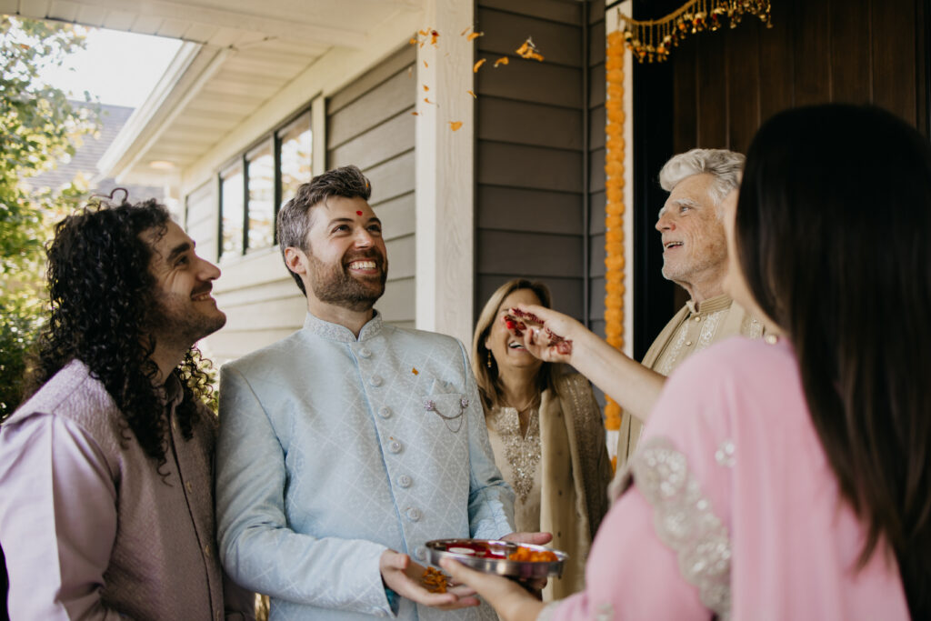 Indian groom in light blue sherwani laughing joyfully during milni ceremony as rose petals are thrown by family at home decorated with marigold garlands, South Asian wedding photography by Mycah Bain Photography Minnesota
