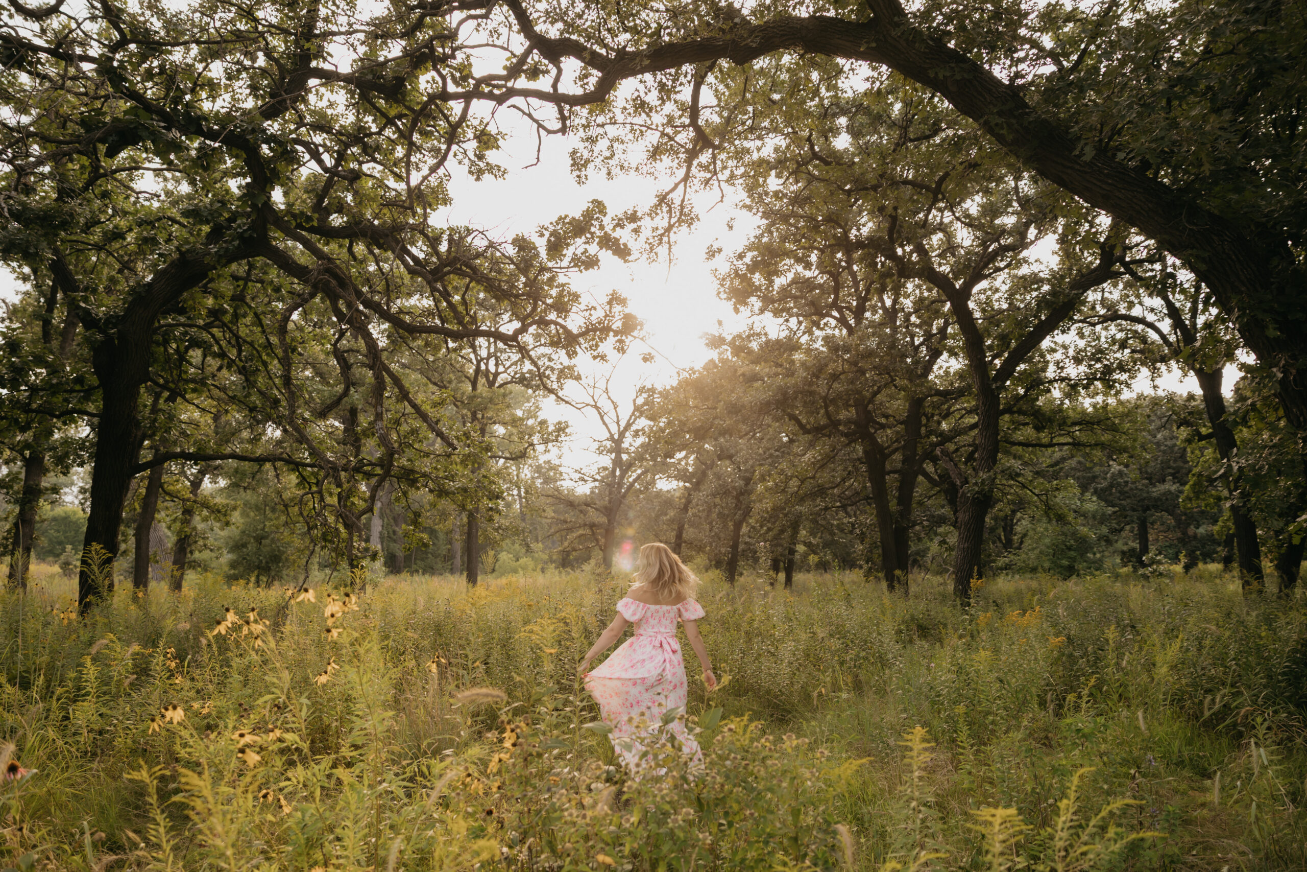 high school senior girl in pink floral dress running through wildflower field at golden hour in Minneapolis