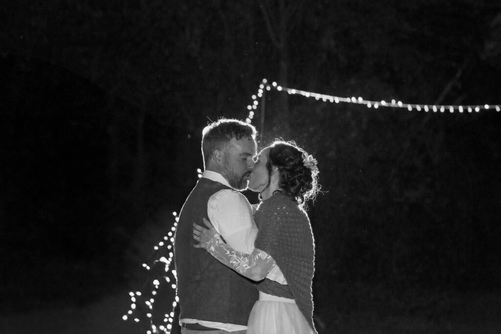 Black and white nighttime photo of bride in lace gown with floral crown and groom in vest kissing under string lights in a dark outdoor setting with rain visible, documentary wedding photography by Mycah Bain Photography Minnesota

