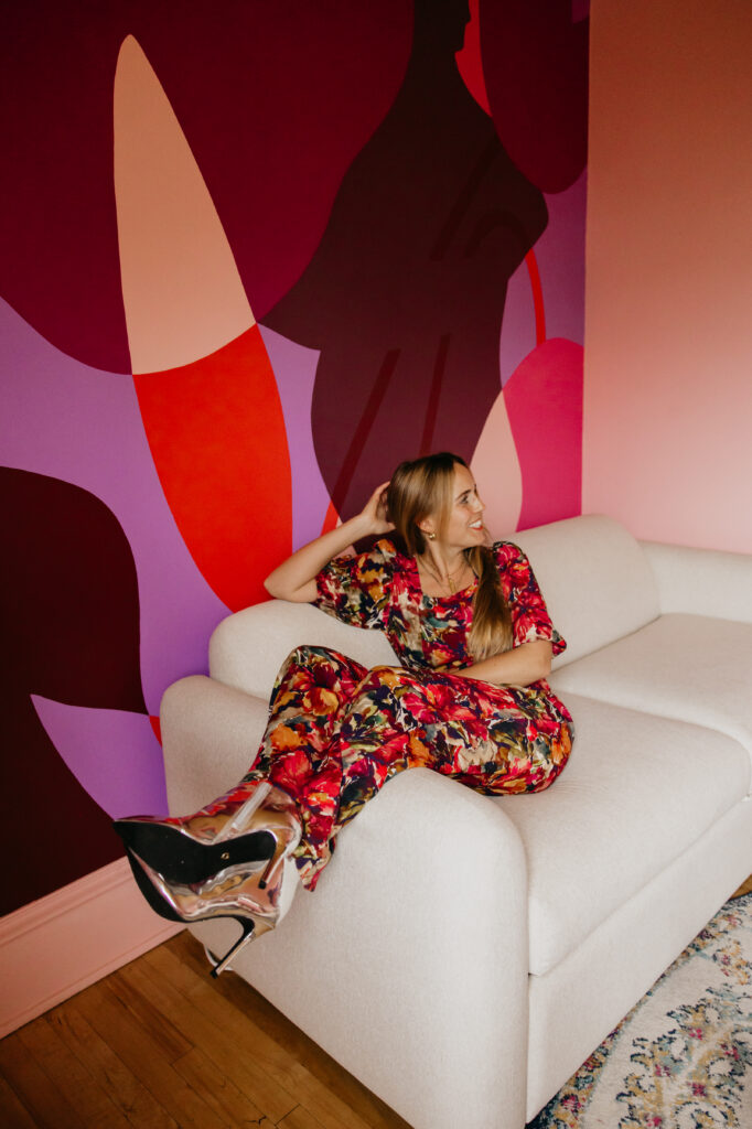 Mycah Bain lounging on a white boucle sofa laughing with her head thrown back, wearing a bold floral jumpsuit and silver pumps in front of a large abstract red and purple mural