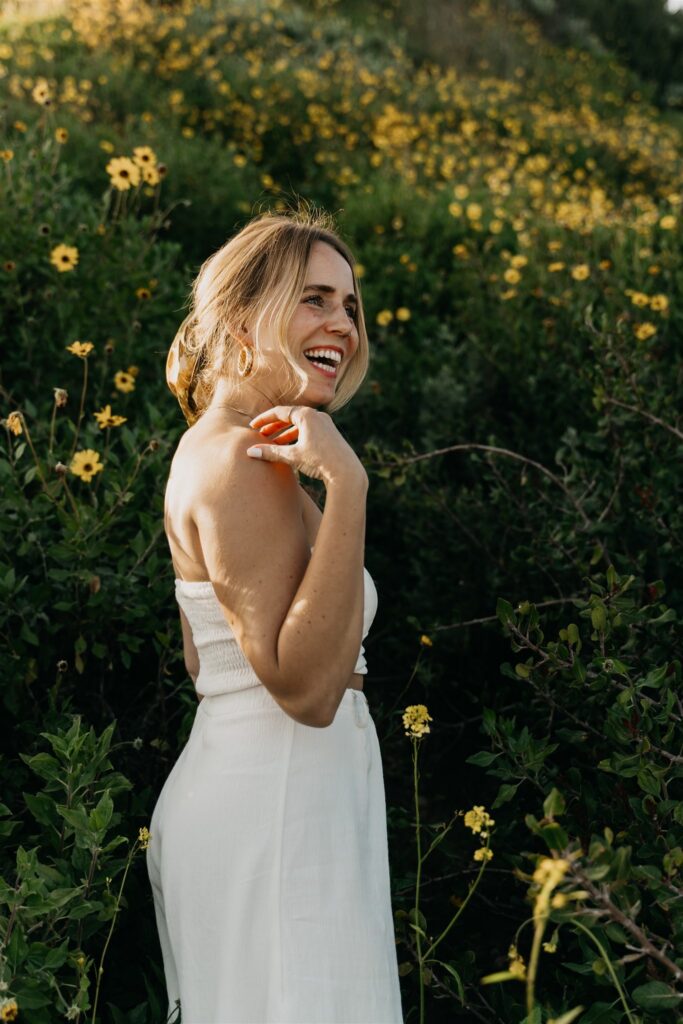 Mycah Bain laughing over her shoulder while standing in a field of yellow wildflowers at golden hour, wearing a white strapless top and wide-leg pants