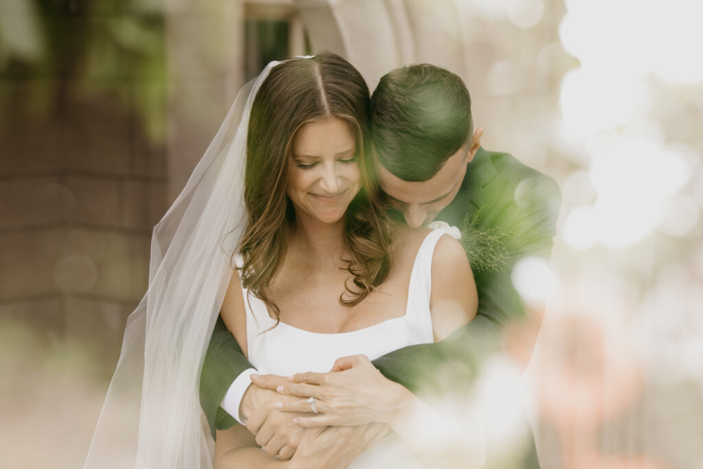 Bride in white square-neck gown smiling softly as groom in olive green suit nuzzles her neck, shot through bokeh foreground, warm golden light, documentary wedding portrait by Mycah Bain Photography Minneapolis