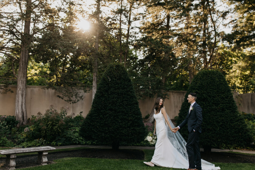 Bride in white slip gown with long veil and groom in navy suit walk hand in hand through a formal garden at sunset with tall pine trees and sunburst in background, documentary wedding photography by Mycah Bain Photography Minneapolis