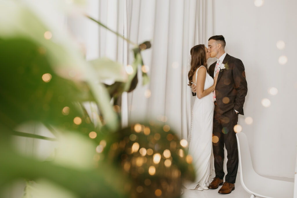 Bride and groom share a kiss against white drape curtains with warm bokeh string lights, lush tropical plant in foreground, wedding at Paris Dining Club Minneapolis, documentary photography by Mycah Bain Photography