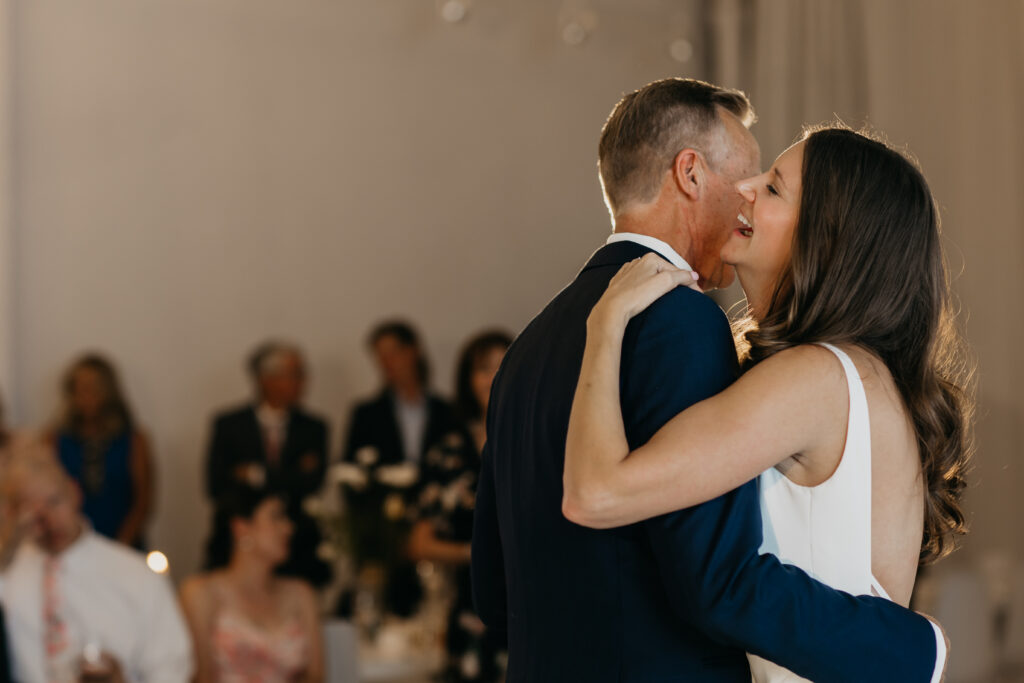 Bride in white square-neck gown and groom in navy suit share first dance, bride laughing and looking up at groom, guests watching in background at Paris Dining Club Minneapolis, documentary wedding photography by Mycah Bain Photography