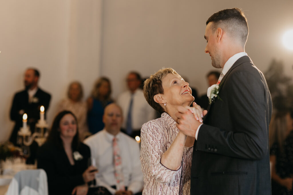 Groom in dark suit dances with his mother in a pink sequined jacket at wedding reception, mother gazing up at him lovingly, candelabras and guests visible in background, documentary wedding photography by Mycah Bain Photography Minneapolis
