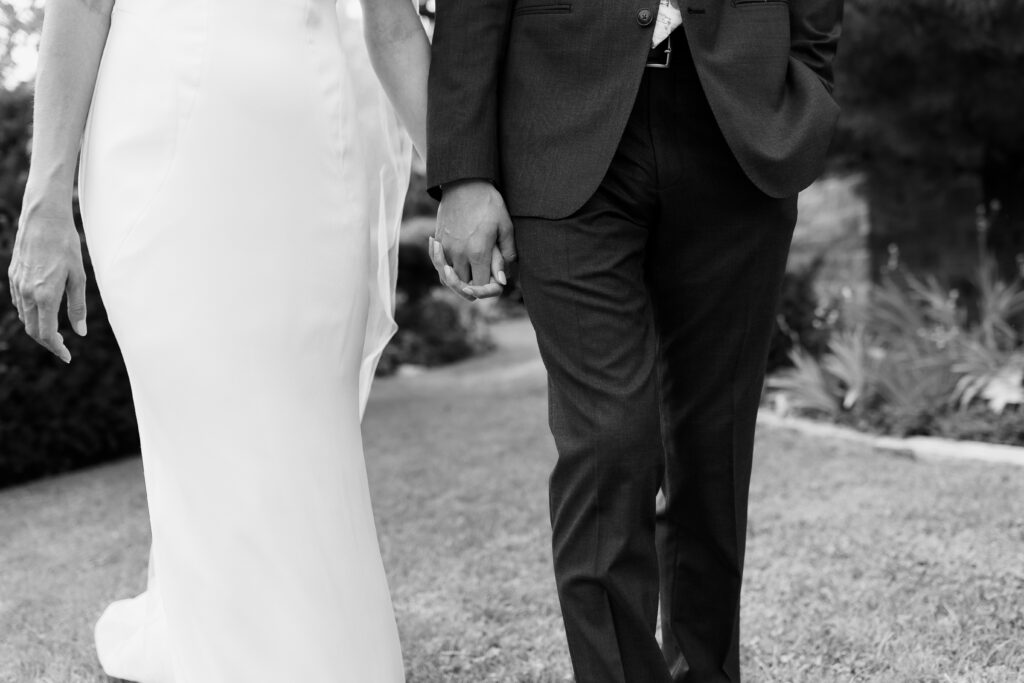 Black and white close-up of bride in sleek white gown and groom in dark suit holding hands while walking outdoors, documentary wedding photography by Mycah Bain Photography Minneapolis