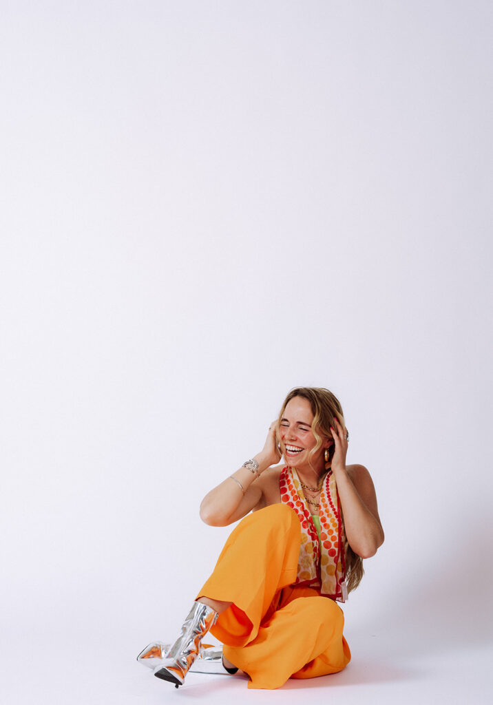 Mycah Bain sitting on a white studio floor laughing with hands on her head, wearing bright orange wide-leg pants, a colorful print top, and silver metallic boots