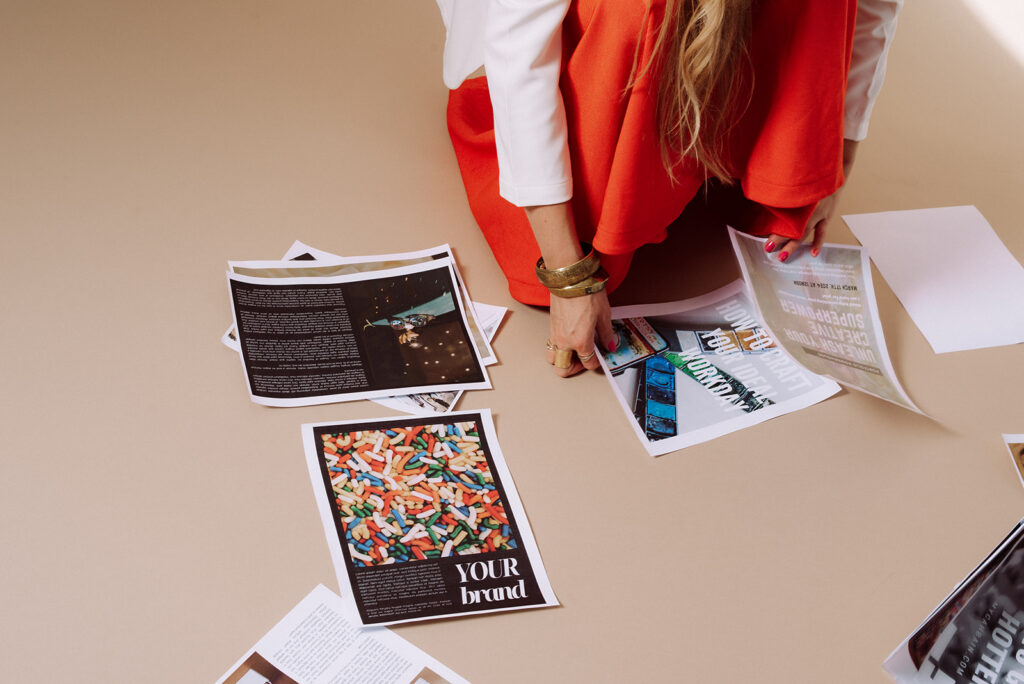 Overhead view of Mycah Bain in a red blazer and white pants kneeling over brand photography print layouts spread across a beige floor, including a piece that reads "YOUR brand"