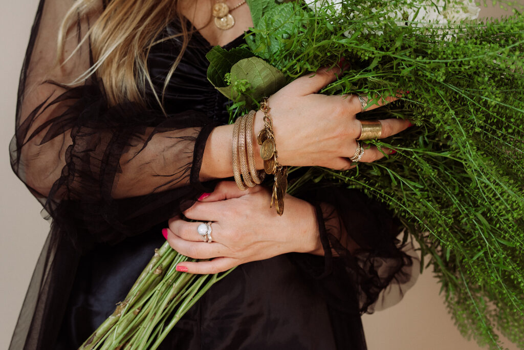 Close-up of hands holding a lush green botanical bouquet, wearing a black sheer ruffled sleeve, layered gold bracelets, a moonstone ring, and a gold cuff
