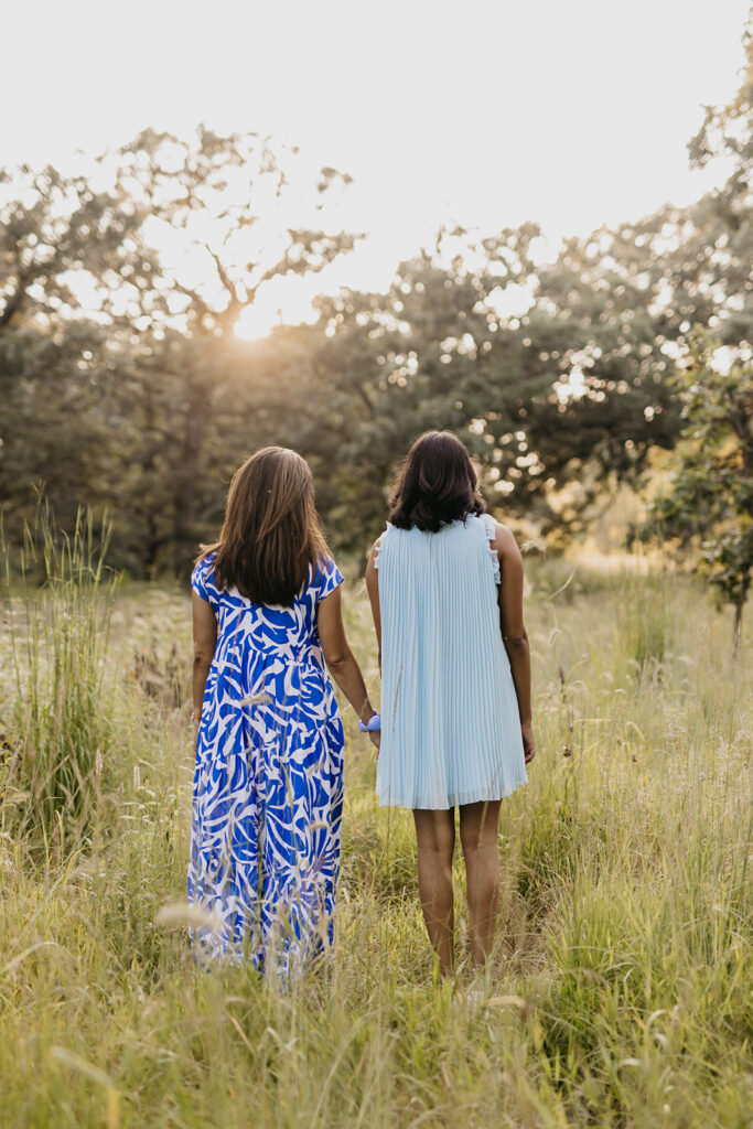 Mom and senior daughter holding hands walking away through golden meadow at sunset senior photo session Minneapolis Minnesota