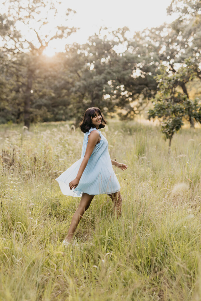 Senior girl in light blue pleated dress walking through golden meadow laughing over shoulder during senior photo session Twin Cities